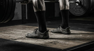 A close-up, low-angle shot of an athlete in a deep squat wearing flat, minimalist barefoot shoes, emphasizing the stable connection between the foot and the gym floor.