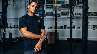 Athlet steht mit seinem Lift More Gym T-Shirt in der Farbe schwarz angelehnt an das Rig und ist bereit für sein Krafttraining